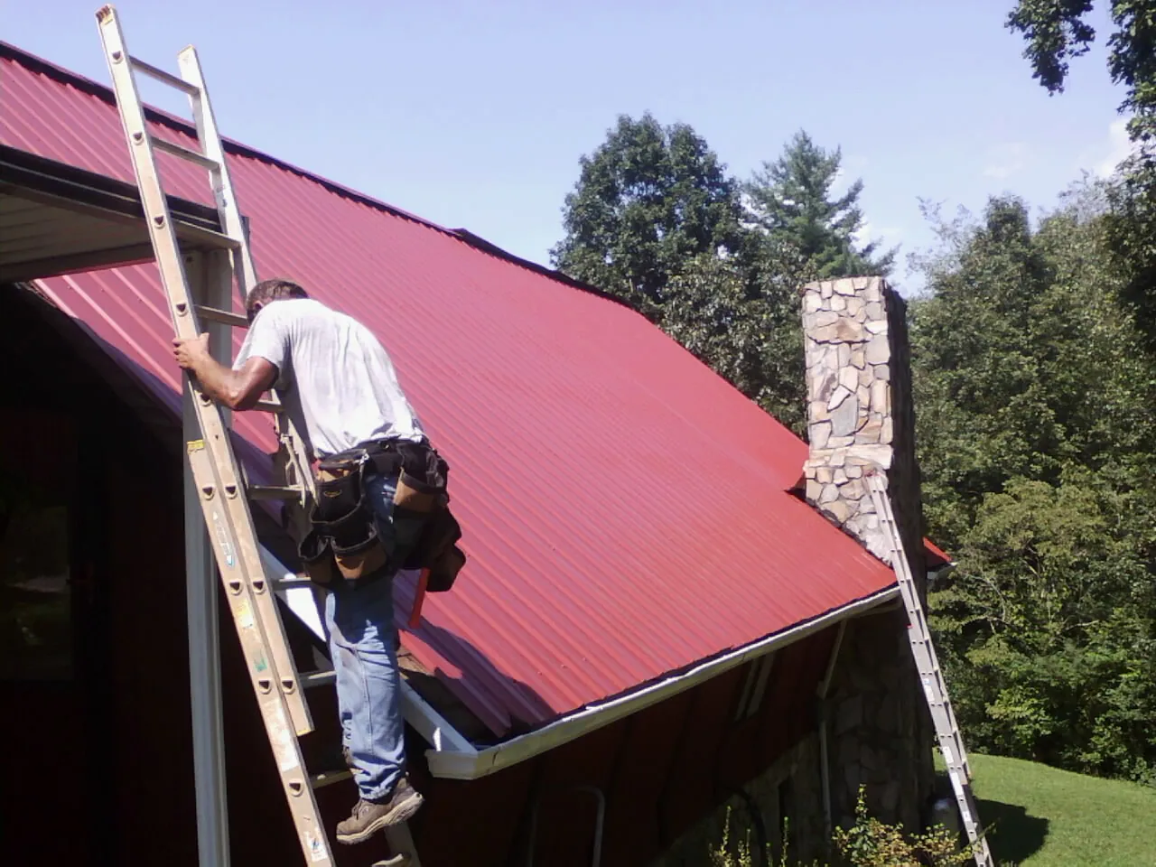 Roofer on a ladder beside a red metal roof and stone chimney — Soffit & Fascia Repair by Roofing Brothers in Manteno